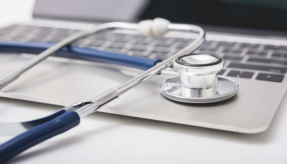 A silver stethoscope with a navy blue tube resting on the keyboard of an open laptop on a white desk