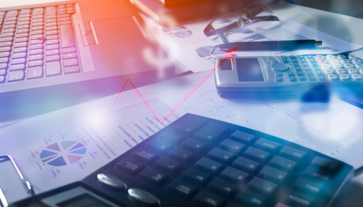 A pen, glasses, two calculators and printed charts on a desk beside a laptop