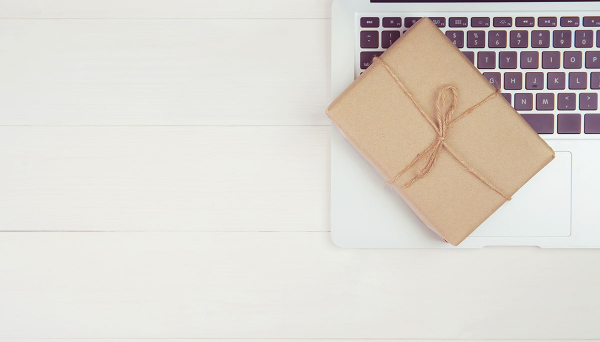 Overhead view of a small gift wrapped in brown kraft paper and tied with twine, resting on the keyboard of an open silver laptop on a white wooden table