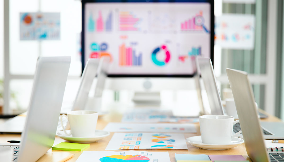 A large monitor showing colourful bar and donut charts at the end of a conference table, with open laptops, white coffee cups, sticky notes and printed charts in the foreground