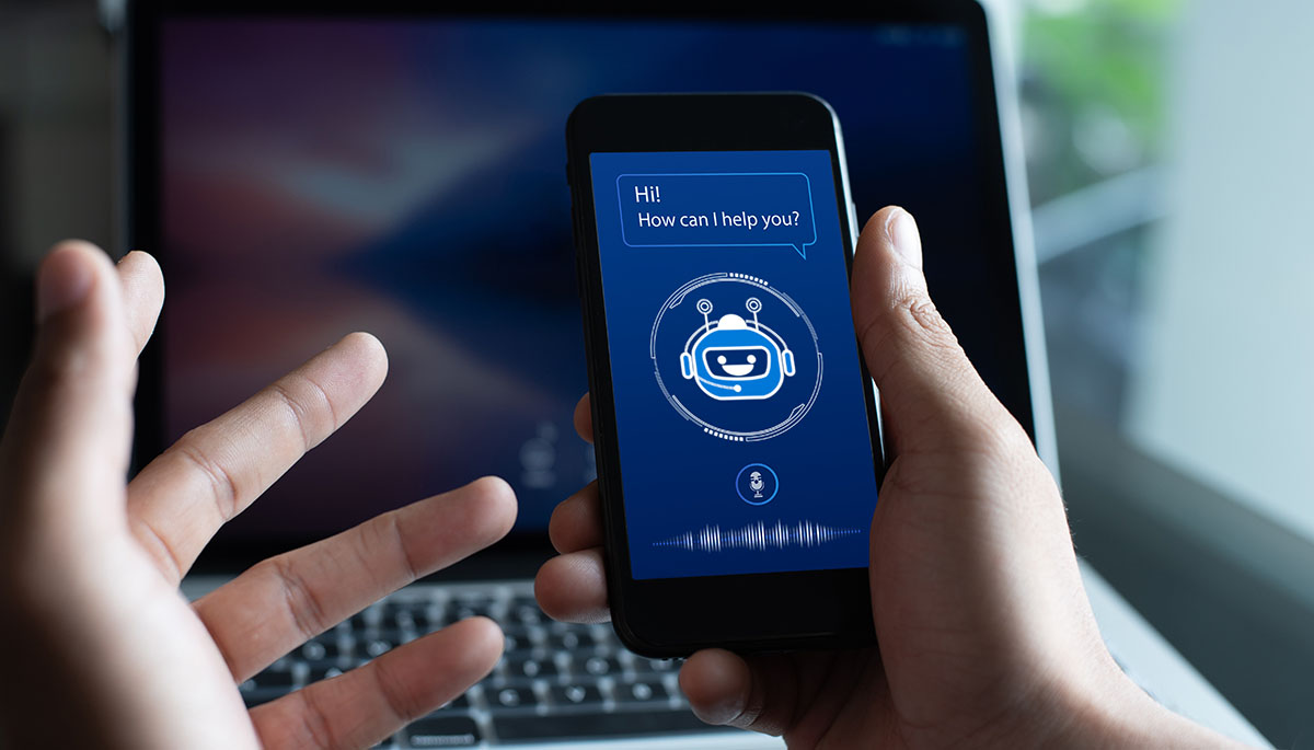 Hands holding a smartphone showing an AI chatbot screen that asks how it can help, in front of an open laptop