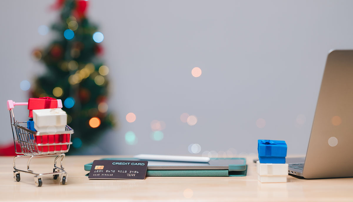 Miniature shopping cart filled with wrapped gifts on a wooden desk beside a credit card, a tablet and an open laptop, with a blurred Christmas tree behind