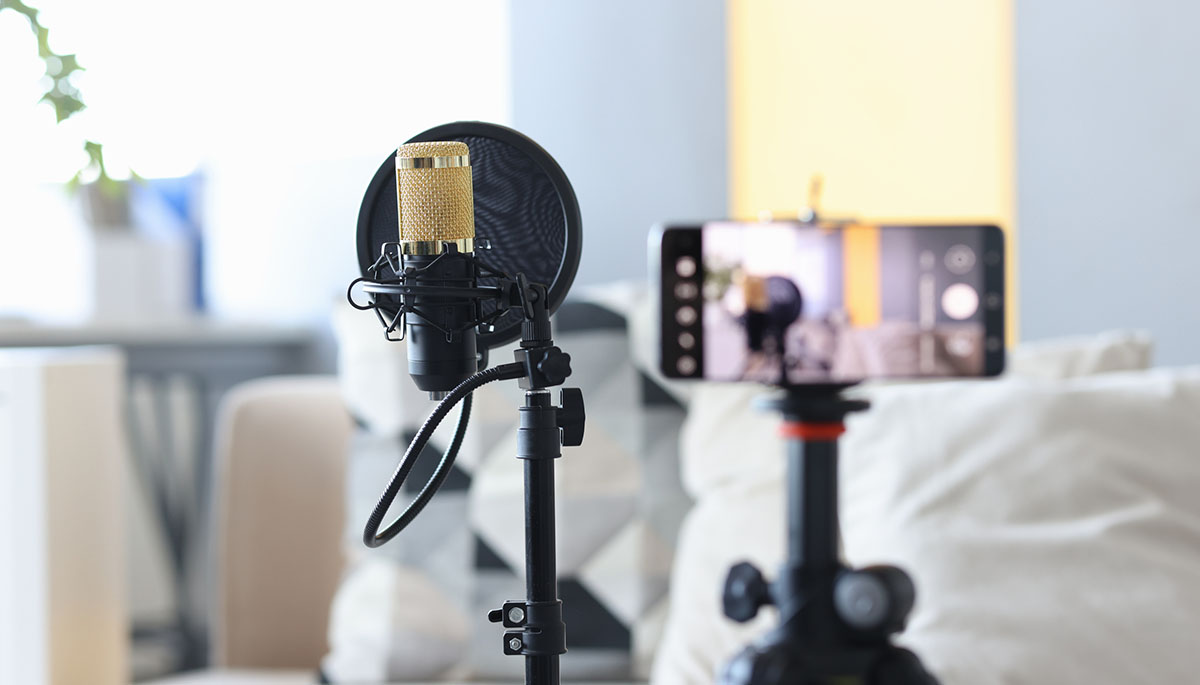 A studio microphone with a pop filter beside a smartphone mounted on a tripod, set up to record video in a bright living room
